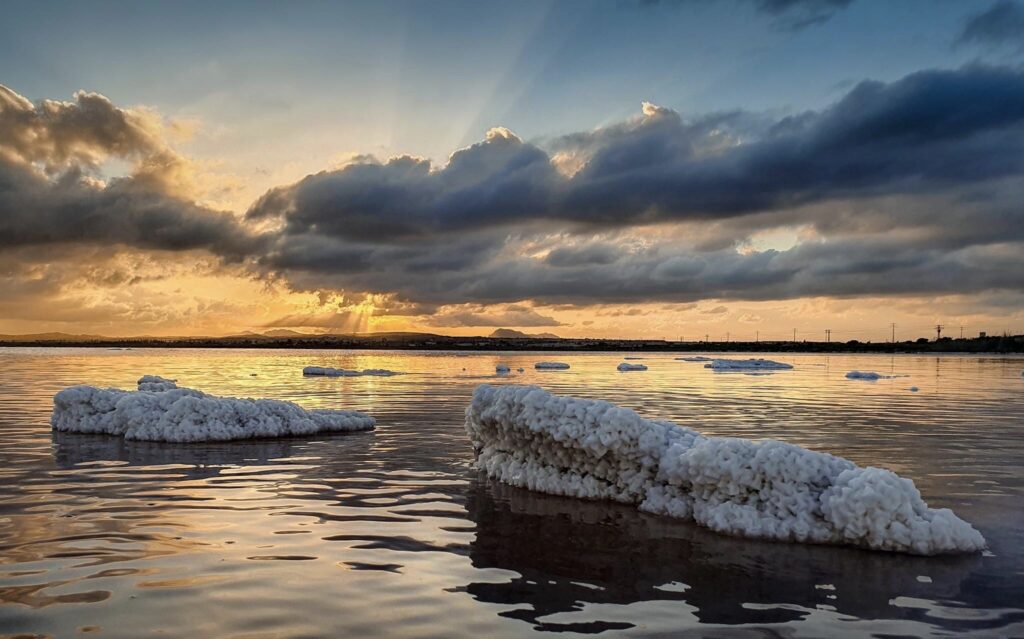 The Pink Lake of Torrevieja glows softly under the warm hues of the setting sun, with the Laguna Salada de Torrevieja reflecting a beautiful tapestry of clouds in its calm, pink waters. The salt crusts along the shore create a striking contrast against the smooth, mirror-like surface of the lake. As the sun dips lower, the colors of the sky—warm oranges and pinks—blend seamlessly with the lake's natural hue, enhancing the ethereal beauty of the scene. The reflections of the fluffy clouds in the Torrevieja salt lake make the water appear endless, with the sky and lake merging into one. The tranquil atmosphere and soft light at sunset bring a serene and almost surreal quality to the pink salt lake of Torrevieja, making it a perfect moment of natural harmony in the Alicante region.