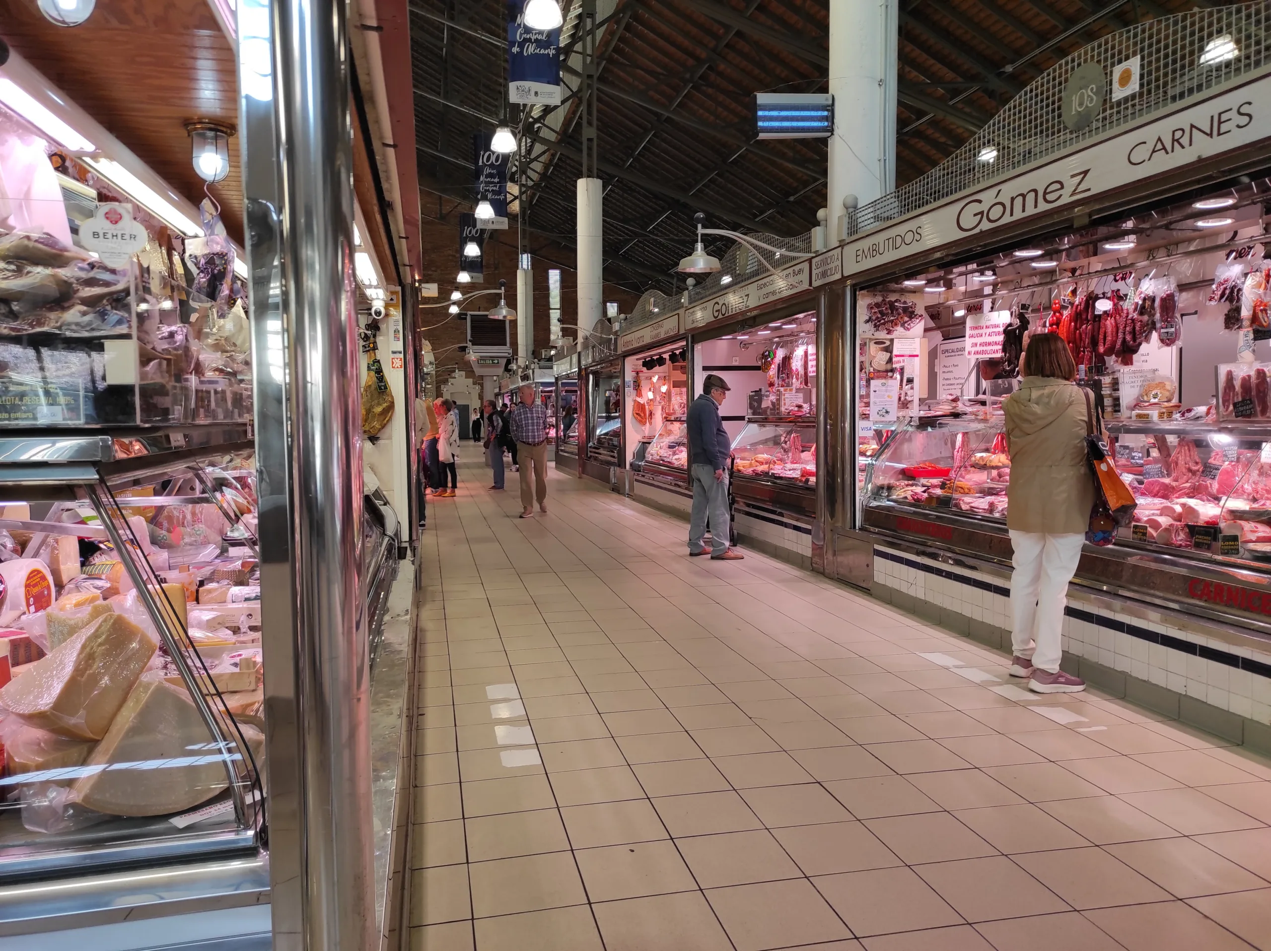 ### Bustling Meat and Cheese Aisle at the Central Market of Alicante

A spacious aisle in the Central Market of Alicante, with various meat and cheese stalls on both sides. Customers are seen browsing and purchasing products, highlighting the market's vibrant atmosphere and wide selection of goods.