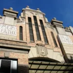 Facade of the Central Market of Alicante, featuring ornate architectural details and mosaic signs reading "Mercado" and "Año 1921" under a clear blue sky.