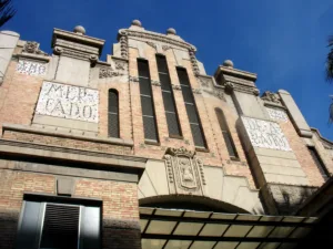 Facade of the Central Market of Alicante, featuring ornate architectural details and mosaic signs reading "Mercado" and "Año 1921" under a clear blue sky.