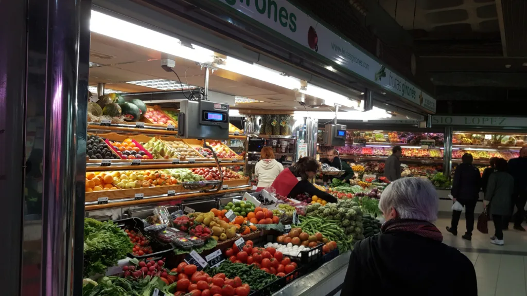 A vibrant fruit and vegetable stall inside the Central Market of Alicante, displaying a colorful and abundant array of fresh produce including bananas, tomatoes, potatoes, and greens, with customers browsing the selection. Mercado Alicante opening hours.