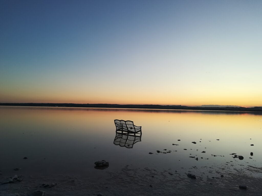 In the heart of the Pink Lake of Torrevieja, a single chair stands on the shimmering surface, its reflection perfectly mirrored in the still, rose-colored waters. The Laguna Salada de Torrevieja, known for its striking pink hue due to the high concentration of salt and microscopic algae, creates a surreal landscape that looks almost otherworldly. The sky above blends seamlessly with the lake, amplifying the dreamlike atmosphere. The contrast between the lone chair and the vastness of the pink salt lake in Alicante evokes a sense of solitude and tranquility. As the sun casts a golden glow over the Torrevieja salt lake, the pink tones become even more vibrant, making this an unforgettable scene in Spain's famous pink lagoon.