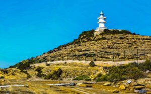 An image taken during the day, with natural sunlight illuminating the lighthouse perched atop a mountain. The lighthouse stands out against the clear blue sky, providing a stunning view of the surrounding nature and mountainous landscape. Cabo de las huertas, alicante, lighthouse