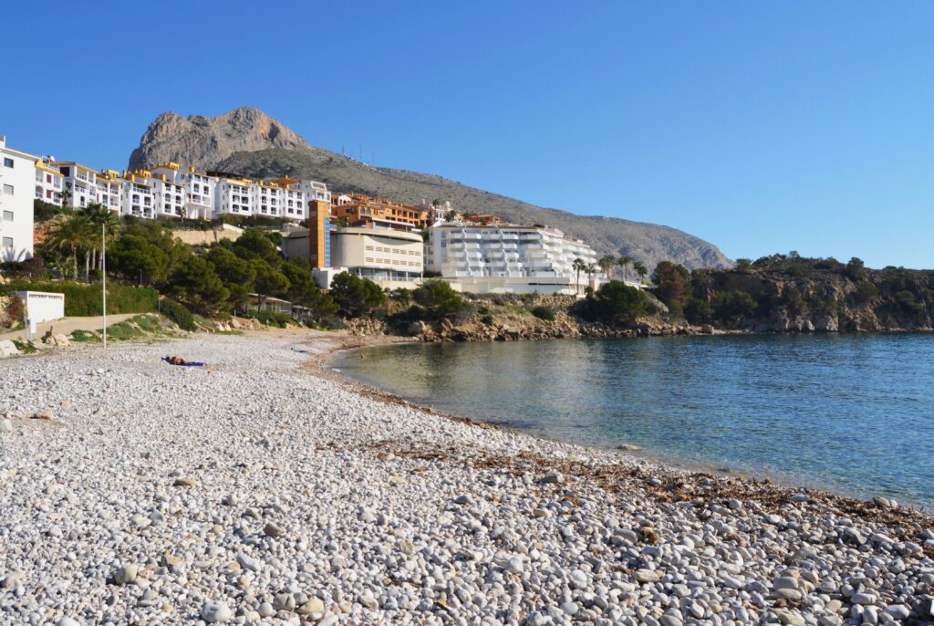 The image depicts a breathtaking panoramic view of Mascarat Beach. The pristine white sand slopes gently towards the crystal-clear turquoise water, creating a mesmerizing contrast. The beach is nestled between dramatic rocky cliffs that rise majestically from the coastline, adding a touch of rugged beauty to the scene.