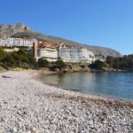 The image depicts a breathtaking panoramic view of Mascarat Beach. The pristine white sand slopes gently towards the crystal-clear turquoise water, creating a mesmerizing contrast. The beach is nestled between dramatic rocky cliffs that rise majestically from the coastline, adding a touch of rugged beauty to the scene.